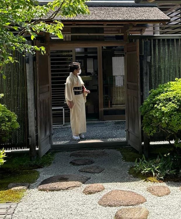 Une femme en kimono clair se tient dans l'encadrement du portail ouvert d'une maison de thé